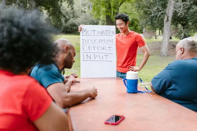 Group of people seated round a table and outdoor listening to a trainer who is holding a card with words scribbled.
